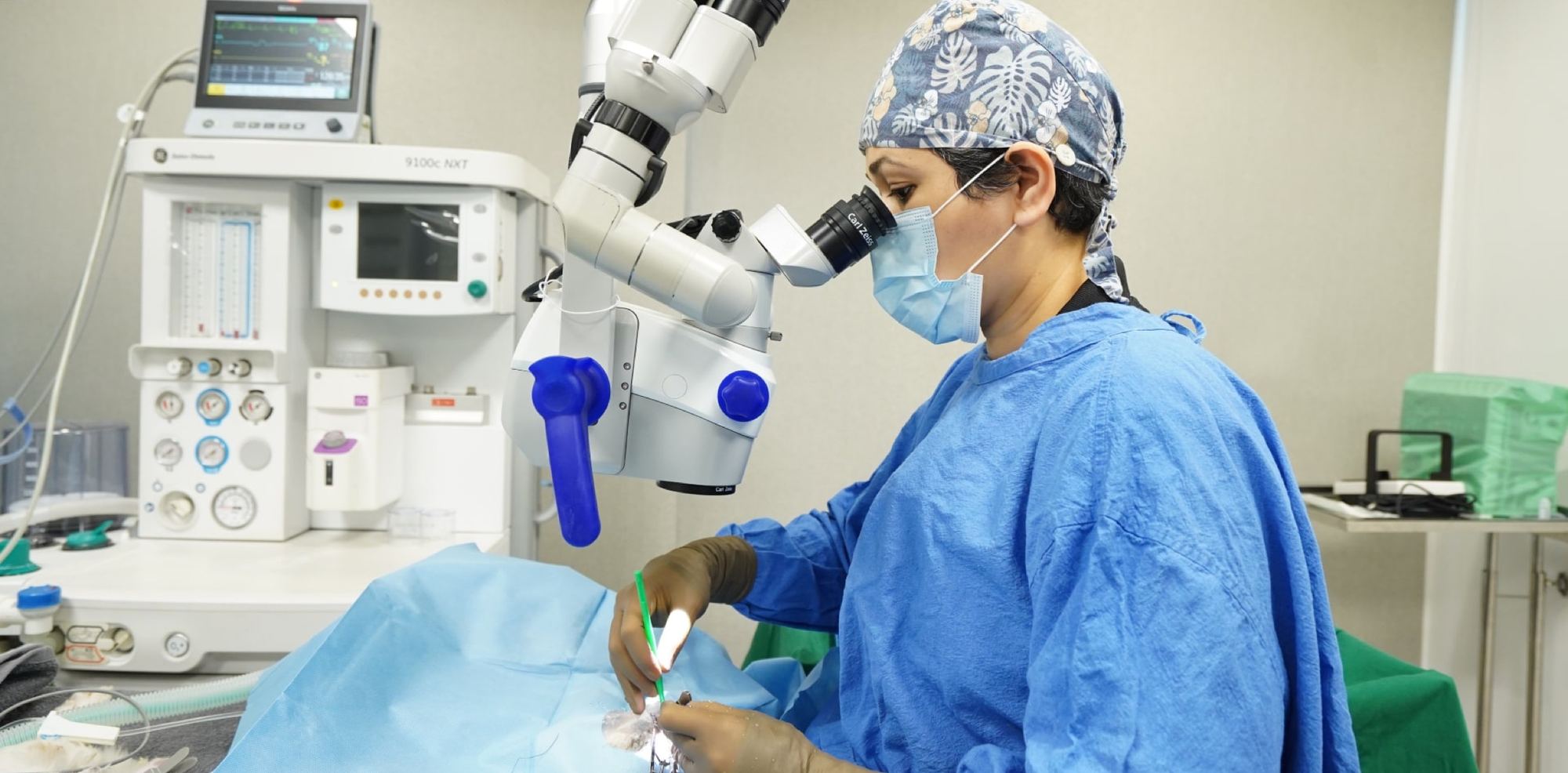 A surgeon in blue scrubs and cap looks through a Carl Zeiss operating microscope over a draped patient, with an anaesthesia machine and monitor behind