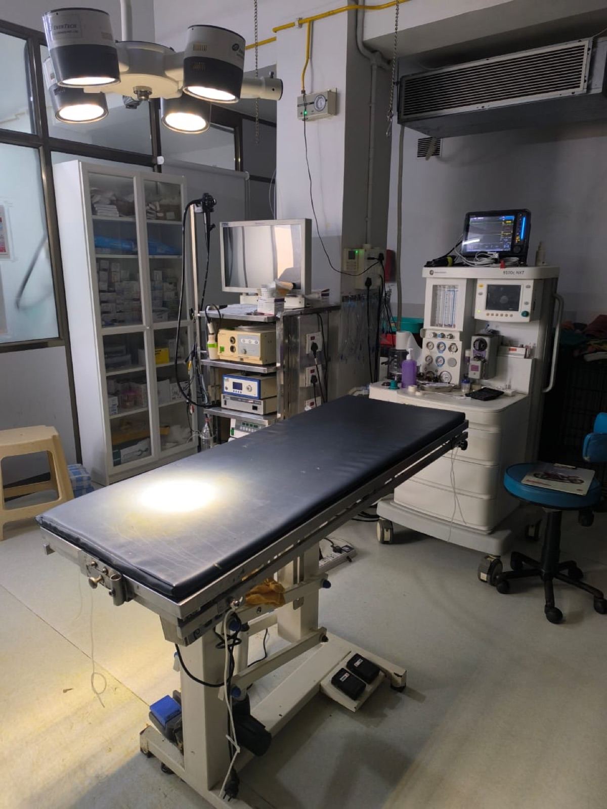 An empty surgical suite with a hydraulic operating table, theatre lights and anaesthesia machine