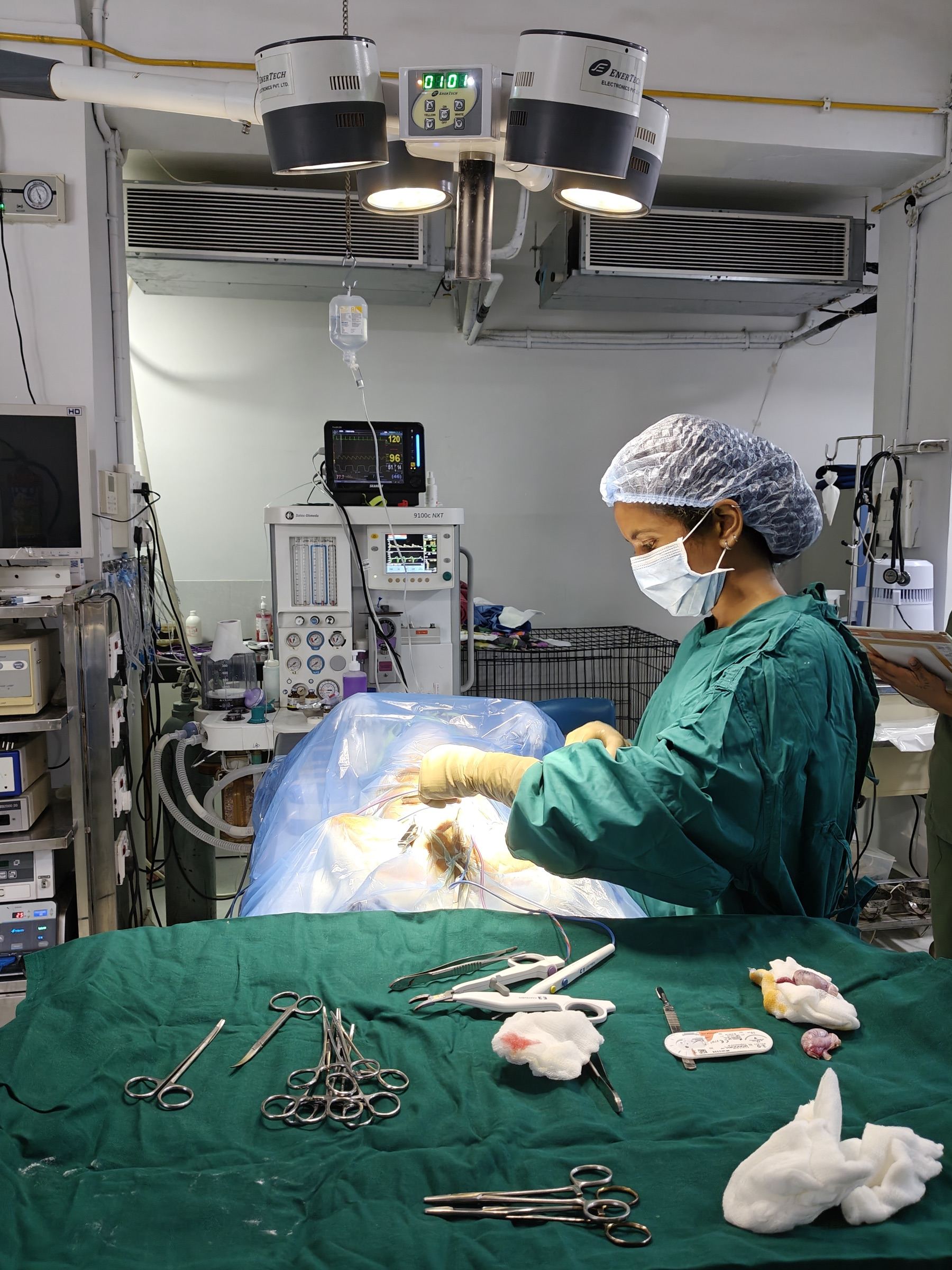 A draped patient on the operating table under twin theatre lights, with anaesthesia machine and monitoring